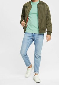 Giacca bomber verde oliva, chiusura con zip, colletto e polsini a coste, indossata su un maglione verde chiaro. Jeans azzurri e sneakers bianche.