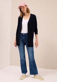 Marineblaue Strickjacke über einem weißen Trägertop, kombiniert mit hellblauen, umflatterten Jeans und beigen Sneakers; zudem wird eine rosa Cordmütze getragen.