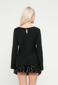 Blusa preta de manga comprida com detalhe em recorte na parte de trás, apresentando uma textura suave e design minimalista, combinada com uma saia preta brilhante de lantejoulas.