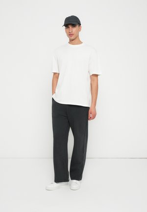 T-shirt blanc à manches courtes associé à un pantalon large gris foncé et des baskets blanches. Une casquette grise achève la tenue, sur fond uni.