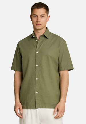 Man wearing a short-sleeve olive green button-up shirt with a subtle horizontal stripe pattern and white pants.