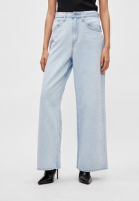 Φαρδιά τζιν παντελόνια ανοιχτού γαλάζιου χρώματος από denim, με ψηλή μέση, μπροστινές τσέπες και αντίθετες ραφές. Φορεμένα με μαύρες γόβες υψηλού τακουνιού.