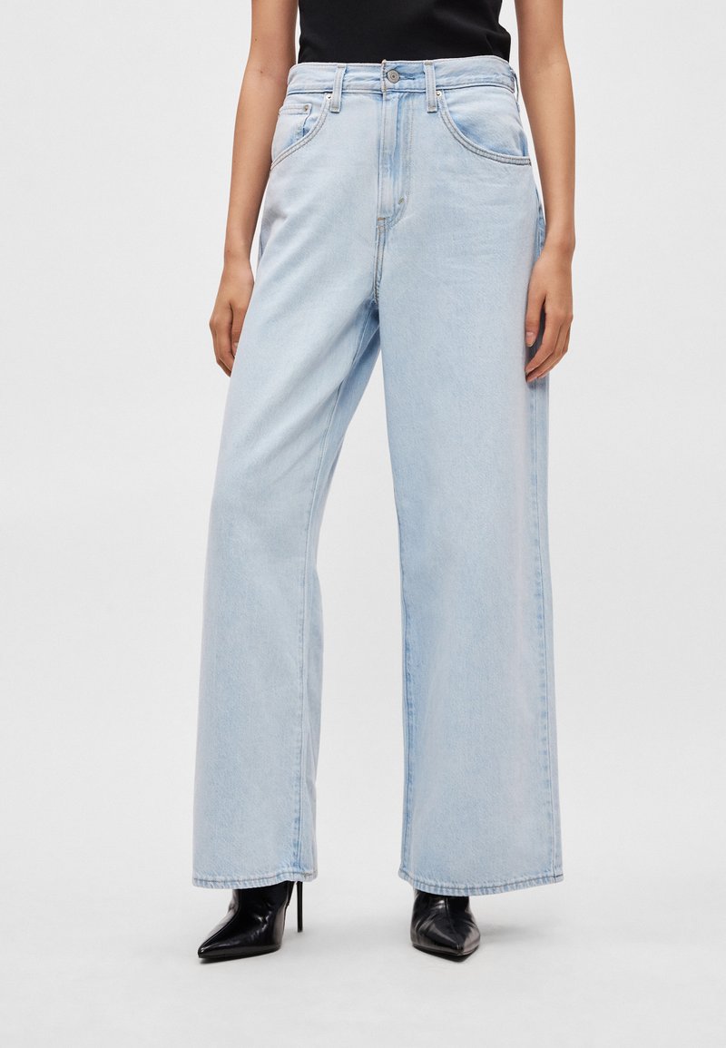 Lyseblå jeans med vide ben lavet af denim, med høj talje, forlommer og kontrastsyninger. Bæres med sorte højhælede sko.