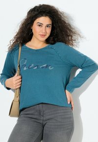 Tyrkysová dlouhá bunda s kulatým výstřihem a textem "POJĎME snít". V kombinaci se šedými slim kalhotami a béžovou kabelkou přes rameno.