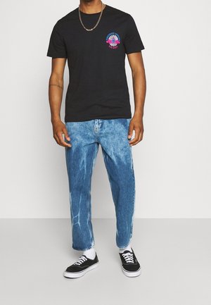 Schwarzes Baumwoll-T-Shirt mit einem bunten Grafikdesign auf der Brust, kombiniert mit hellblauen Jeans und schwarzen Leinenschuhen.