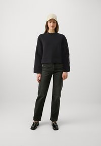 Afgeknotte zwarte sweatshirt met brede mouwen, gecombineerd met donkere spijkerbroek en glanzende zwarte schoenen. Beige pet, minimalistisch logo detail op de mouw.