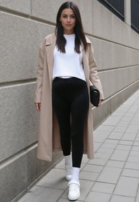 Bežový dlouhý kabát přes bílý crop top, kombinovaný s černými legínami a bílými teniskami. Oblečení se vyznačuje čistými liniemi a minimalistickým designem.