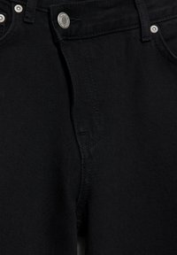 Detailní záběr černých džínů z denimu zobrazující kovové knoflíky, zip, poutka na pásek a švy na předních kapsách.