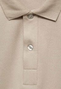 Beige pikétröja i strukturelltyg, med ribbad krage, knappslå med två knappar och en liten fickdetalj under knapparna.