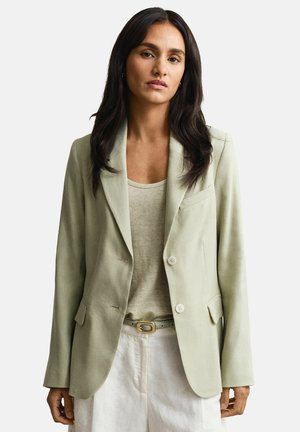 Frau mit langen dunklen Haaren trägt ein hellbeiges Sakko über einem passenden Oberteil und weiße Hose mit beigem Gürtel, vor schlichtem weißem Hintergrund.