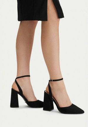 Kobieta nosząca czarne czółenka z odkrytym piętą, z paskami wokół kostki, na stabilnym obcasie i o szpiczastym nosku oraz czarną spódnicę z bocznym rozcięciem.