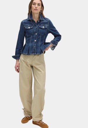 Frau trägt eine eng anliegende dunkelblaue Jeansjacke mit ausgestelltem Saum, beige weite Hosen mit Schnürung am Knöchel und braune Mokassins, steht mit der Hand in der Hüfte.