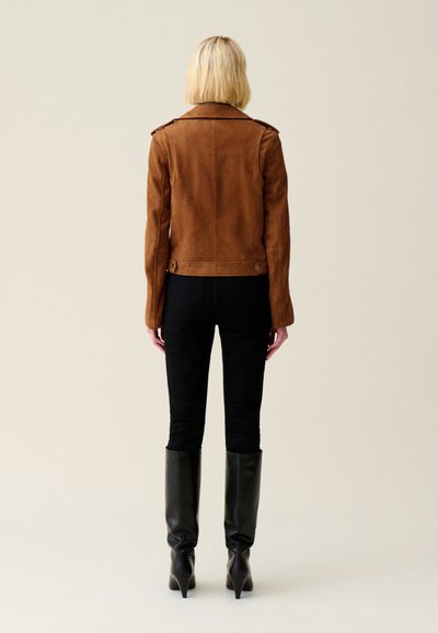 Giacca da moto in suede marrone con spalline, dettaglio con cordoncino sul retro, abbinata a pantaloni neri aderenti e stivali neri con tacco alti fino al ginocchio.