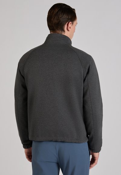 Mężczyzna widziany od tyłu, ubrany w ciemnoszarą kurtkę zapinaną na zamek z elastycznymi mankietami oraz niebieskie spodnie na jednolitym, jasnym tle.