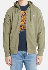 Personne portant un sweat à capuche zippé vert olive par-dessus un t-shirt graphique bleu marine et un jean bleu clair, debout avec les bras détendus le long du corps.