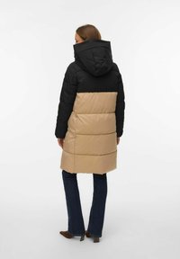 Pufota mētelis ar melnu augšējo daļu un bēša apakšējo daļu. Iekļauj kapuci un vertikālu šuvumu. Valkāts virs tumšām platiem džinsiem.