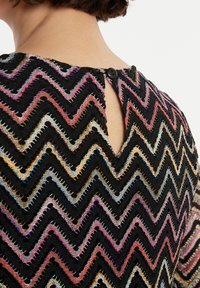 Top en tricot avec un motif en chevron noir réalisé en fils multicolores, avec un détail de dos en forme de trou de serrure et une fermeture à un bouton.