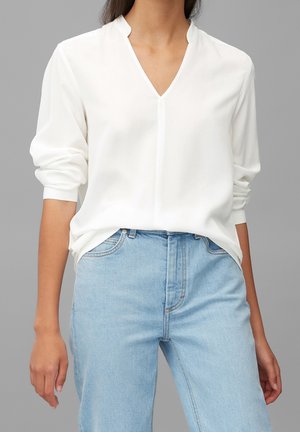 Personne portant un chemisier blanc à manches longues avec une encolure fendue et un jean bleu clair taille haute sur fond gris.