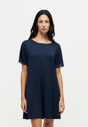 Kvinde iført en løs navyblå kjole med korte ærmer og blonder på ærmerne, stående mod en ensartet hvid baggrund.