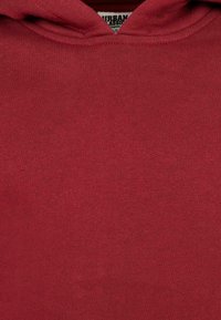 Nahaufnahme eines roten Hoodies, die die Textur und die Kapuzennaht zeigt, mit einem teilweise sichtbaren "Urban Classic"-Etikett im Kragen.