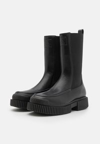Mustad nahast mustad pahkluu saapad elastsete külgpaneelidega, paksude ribaliselt mustritega taldadega ja esiosas embosseeritud logoga.