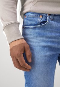 Gros plan sur la main d'une personne reposant près de la poche d'un jean en denim bleu, portant un haut beige à manches longues.