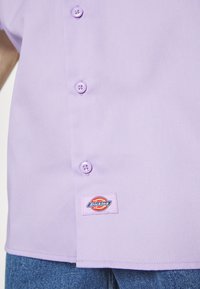 Lavendel kortärmad skjorta med en mjuk textur, prydd med lätta knappar och en Dickies-etikett på den nedre vänstra sidan.