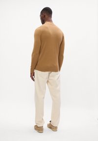 Maglione lungo aderente marrone con collo rotondo, abbinato a pantaloni beige chiari e scarpe marroni slip-on, visto da dietro.