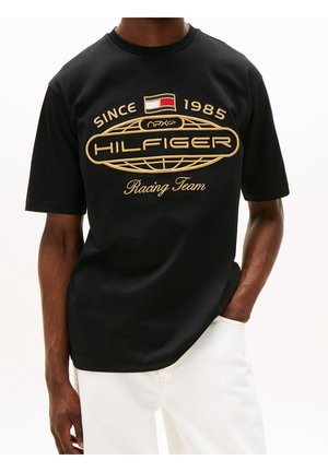Person mit schwarzem Hilfiger Racing Team T-Shirt mit goldenem Logo-Schriftzug und weißer Hose, steht mit einer Hand in der Tasche.