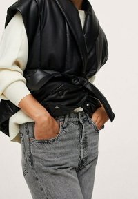 Chaqueta sin mangas de cuero negro con cintura con cinturón, combinada con jeans de cintura alta grises, que presentan bolsillos frontales y un ligero efecto de lavado.