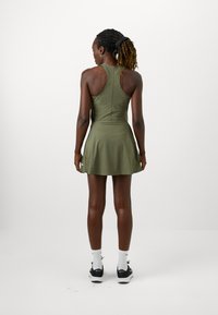 Vestido de tenis elástico en verde oliva con falda acampanada, diseño de tirantes tipo racerback y un pequeño logo en el dobladillo. Textura suave, longitud hasta medio muslo.