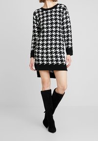 Černobílé pletené šaty v houndstooth vzoru s dlouhými rukávy, kulatým výstřihem a asymetrickým lemem, doplněné černými zamíchanými kozačkami po kolena.
