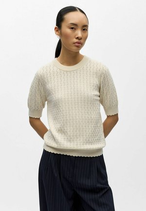 Kvinne med mørkt hår i hestehale som har på seg en kremfarget strikket kortermet genser og mørke bukser med striper, stående med hendene bak ryggen.