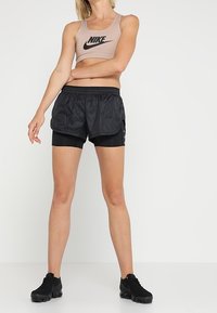 Ljus beige Nike sport-bh med svart logga, i kombination med svarta löpshorts med lagerdesign. Modellen bär svarta träningsskor.