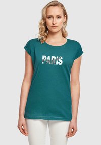 Türkises T-Shirt mit kurzen Ärmeln, auf dem das Wort "PARIS" in fetten weißen Buchstaben mit grafischer Textur zu sehen ist, kombiniert mit weißen Jeans.