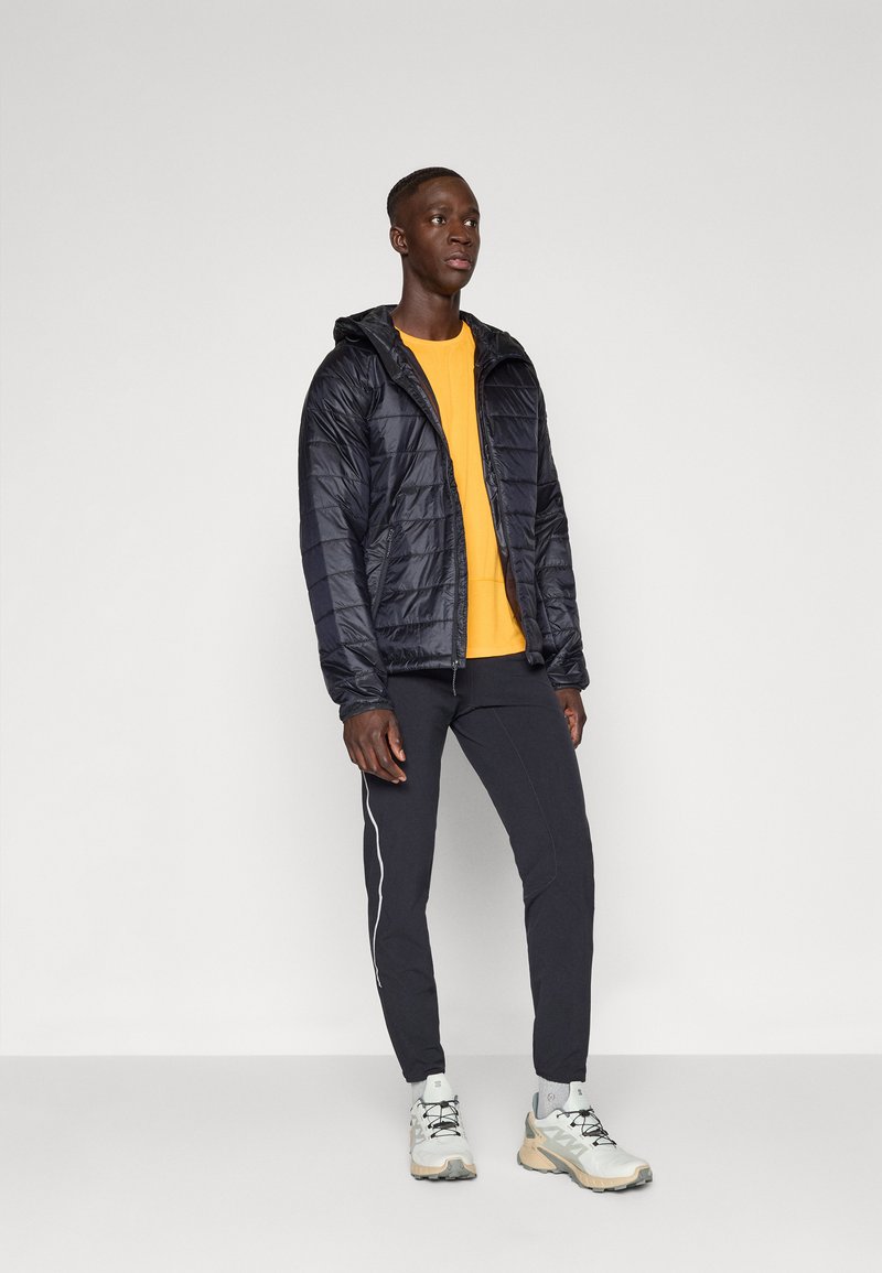 Jeune homme portant une doudoune noire par-dessus une chemise jaune, un pantalon de sport noir et des baskets blanches, se tenant devant un fond blanc uni.