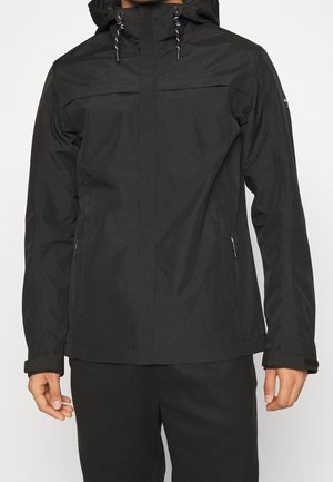 Mann trägt eine schwarze Kapuzenjacke mit Reißverschlusstaschen und verstellbaren Bündchen, steht vor einem einfarbigen hellen Hintergrund.