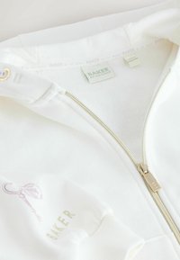 Valkoinen vetoketjullinen collegepaita, jossa on kultaista vetoketjua, pehmeää kangasta ja minimalistiset violetit grafiikat, joissa on avain ja sana "BAKER" hihassa.