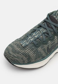 Scarpa sportiva a motivo verde e beige con tomaia in tessuto testurizzato, sistema di allacciatura strutturato e logo Nike nero sul lato.