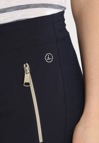 Los pantalones azul marino cuentan con una cremallera lateral con herrajes dorados y un logo circular realzado por pequeños cristales de imitación. El tejido tiene una textura suave.