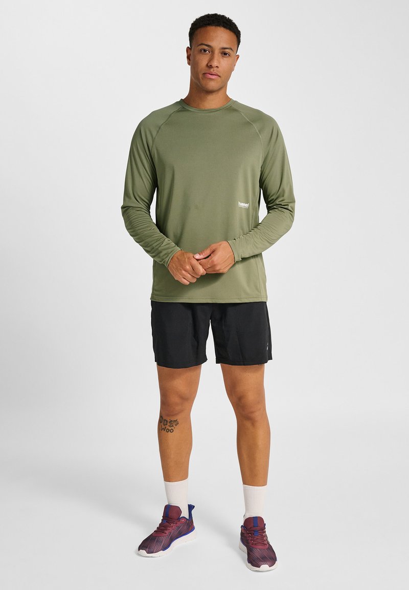 Mand stående iført olivengrøn langærmet skjorte, sorte shorts, hvide sokker og lilla løbesko mod en ensfarvet lys baggrund.