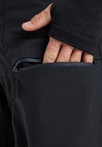Zwarte softshell broek met een ritszak. Een close-up toont een hand die in de zak reikt, met nadruk op de materiaal- en stikdetails.
