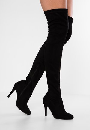 Jambes portant des cuissardes en daim noir avec des fermetures éclair latérales et des bouts pointus sur des talons aiguilles hauts sur fond blanc.