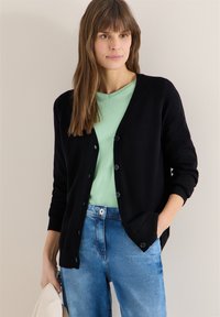 Femme portant un cardigan noir boutonné sur une chemise vert menthe et un jean bleu, tenant un chapeau beige dans une main, debout contre un mur uni.