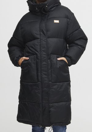 Longue doudoune noire avec capuche, fermeture éclair à l'avant, deux grandes poches avant, poignets élastiques et une petite écusson logo sur la poitrine. Mains dans les poches.