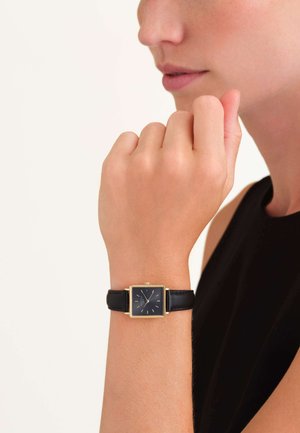 Orologio quadrato nero con cinturino in pelle nera e quadrante semplice, con una cornice dorata. La mano del modello poggia vicino al mento, indossando un top nero.