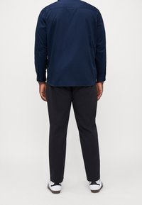 Camicetta a maniche lunghe blu scuro con polsini abbottonati, abbinata a pantaloni grigi scuri e sneaker bianche con accenti neri.