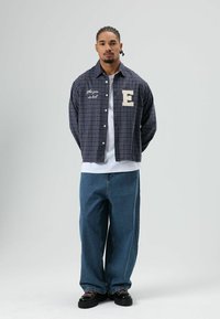 Geruit overhemd in donkerblauw met witte accenten; voorzien van knopen, een geborduurde letter 'E' en tekst; gecombineerd met losse blauwe jeans en zwarte schoenen.