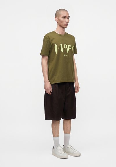Άνδρας που φοράει ελιάς πράσινο t-shirt με το κείμενο «Hope», μαύρα σορτς μέχρι το γόνατο, λευκές κάλτσες και λευκά αθλητικά παπούτσια, στέκεται μπροστά σε απλό φόντο.