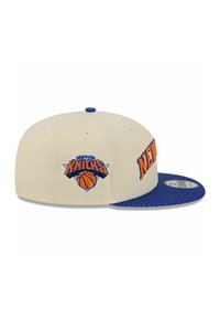 Light beige baseball cap with a blue brim, featuring embroidered "New York Knicks" logo in orange, and a basketball graphic.